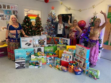 Belly dancers from Sparkles Belly Dance classes with gifts they donated. Image shows three belly dancers in costume and one member of staff, not in clinical uniform, with a Christmas tree and toys. They are doing a belly dance pose with their arms.