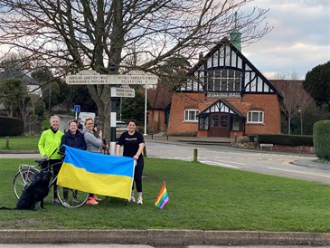 The four friends who are undertaking a charity challenge for Ukraine. Image shows four women standing on a green area in front of a village hall, by a sign. They are holding a Ukrainian flag. There is also a rainbow LGBT flag on the ground. One of th