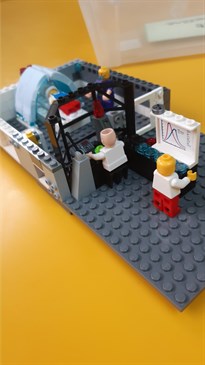A lego model of the MRI scanning room. The control room can be seen with various lego figures as medical staff, plus the scanning room with scanner and a lego patient