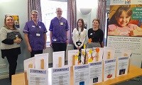 Avatar of the research team at the coffee morning. Image shows a group of people standing behind a table, which has signs about different research studies and a small tree in the centre with coloured paper 'leaves' hung from it. There is also a banne