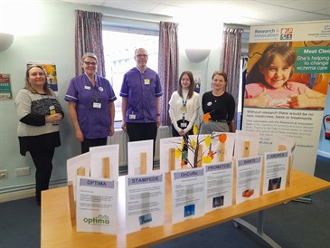 The research team at the coffee morning. Image shows a group of people standing behind a table, which has signs about different research studies and a small tree in the centre with coloured paper 'leaves' hung from it. There is also a banner promotin