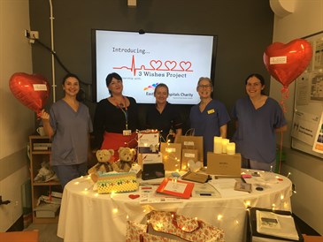 The team at the Kent and Canterbury Hospital at the launch of the 3 Wishes Project there. Image shows a group of staff standing behind a table with various items on. Behind them is a screen that says introdcing the 3 Wishes Project
