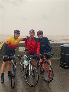 The team battled poor weather conditions to make the finish in time. Image shows three of the four men with bikes on a seafront in bad weather.
