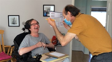 Tony Wright, in a wheelchair, with carer Richard Wood. Richard is holding the Eye Talk device and using it to help Tony communicate
