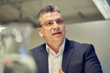 Head and shoulders of Zach Tsiamoulos, the Trust's lead for endoscopy. He is speaking at an event. There is a jug of water out of focus in the left foreground of the image