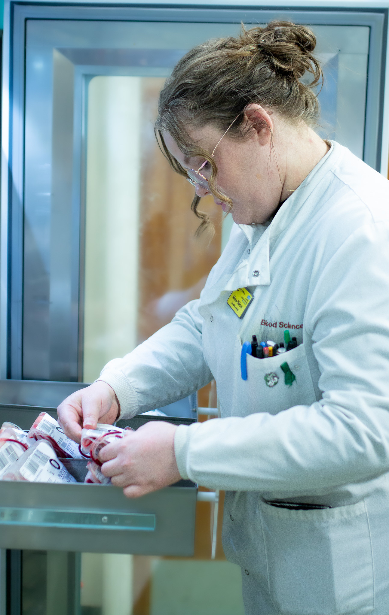 Allana Dickson. She is pictured removing a blood bag from a fridge, wearing a white lab coat and yellow name badge