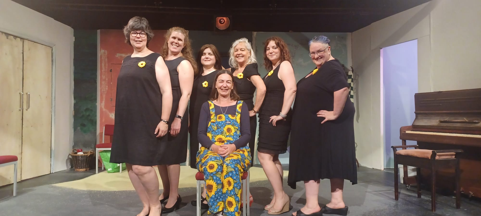 Andrea Oliver and the six main cast members of the production of Calendar Girls at the Arden Theatre in Faversham. Adrea is sitting on a chair wearing dungarees made from a sunflower material, and the six women are standing behind her wearing black dresses with crochet sunflower brooches