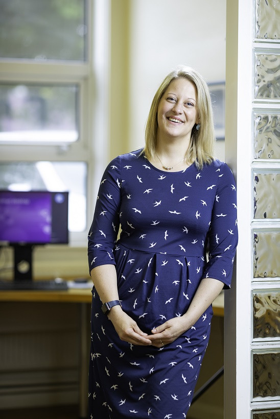 Carly Larkin, who will take on a wing walk for East Kent Hospitals charity. She is leaning on a glass tiled wall in an office wearing a blue dress.