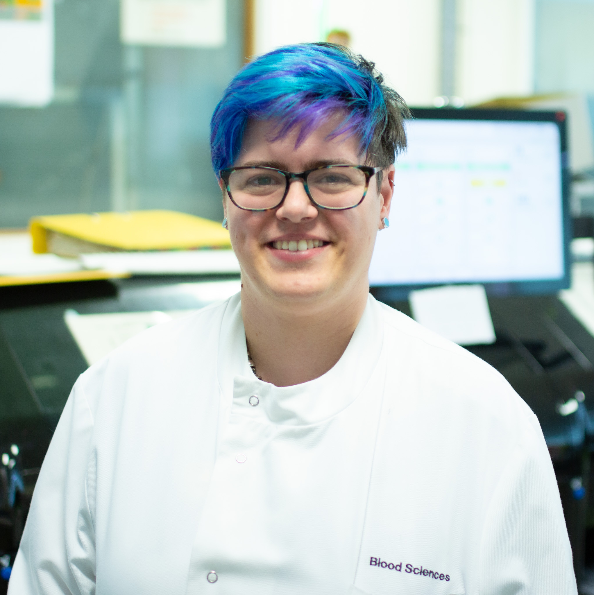 Hannah Scholes, biomedical scientist. Image shows Hannah wearing a white lab coat with blood sciences written on it, with an analyser behind her