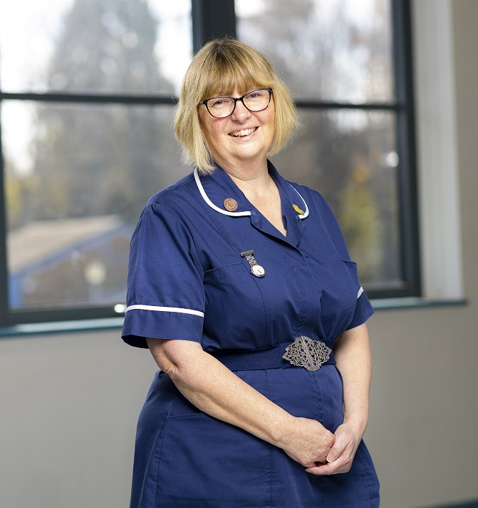 Karen Brewer - image shows her standing in front of a window in a dark blue nurse's uniform with belt