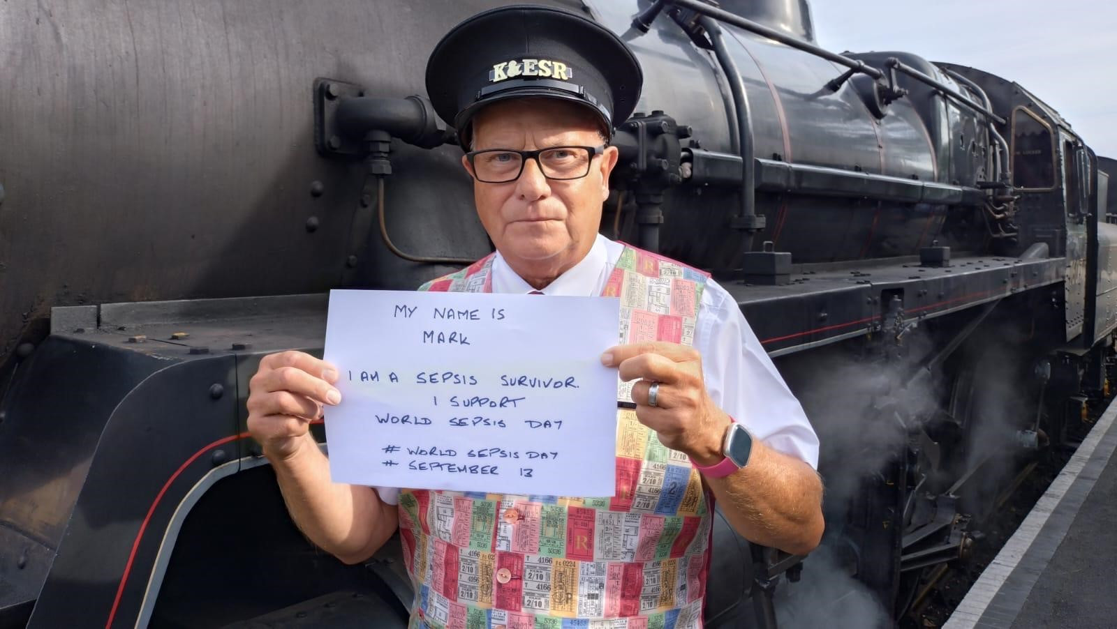 Mark Wignall, holding a sign with his name, and that he is a sepsis survivor and supports world sepsis day. He is wearing a train drivers hat and is in front of a steam train