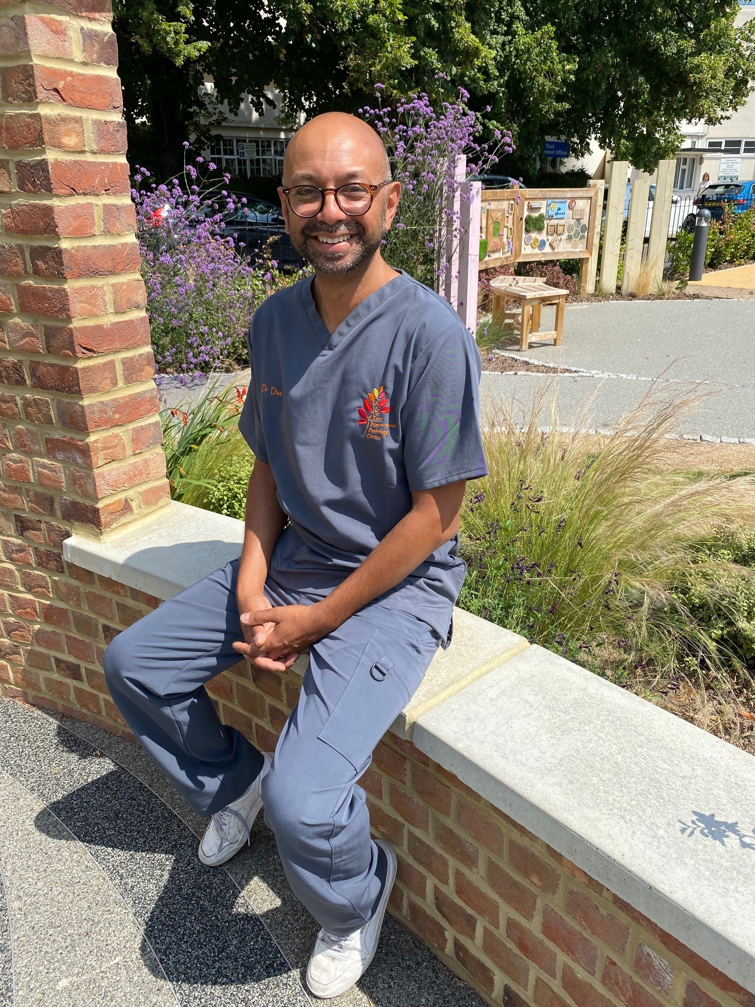 Dr Neelan Das, Trust AI lead and consultant radiologist. He is pictured sitting on a low wall in a garden wearing scrubs