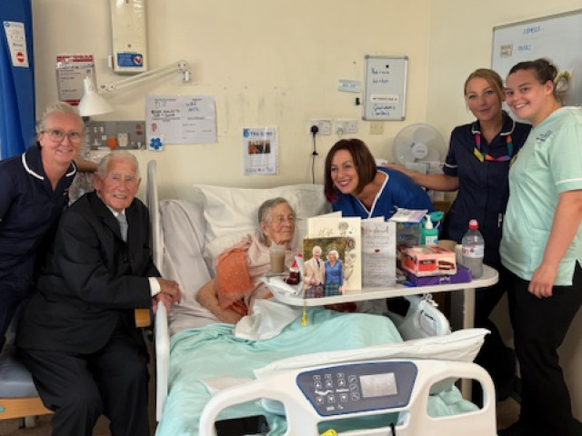 Pat and Jock celebrate their anniversary with staff at the QEQM. Pat is in a hospital bed with cards on the table in front of her