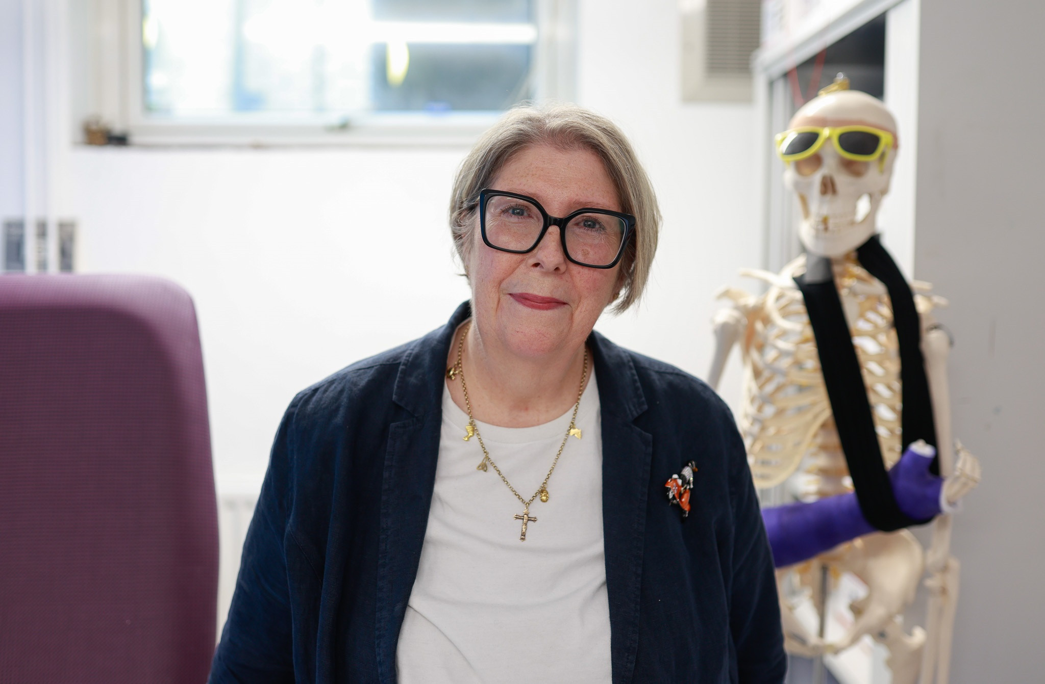 Shirley Bowskill. Image shows her in a clinical room with skeleton with arm in plaster and sunglasses behind