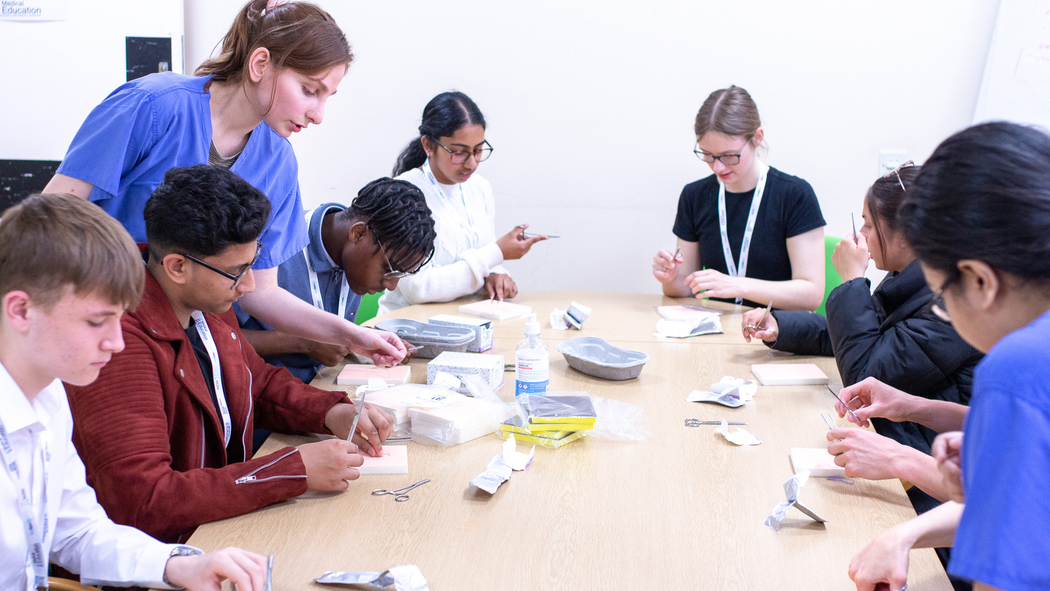 Students practising suturing at the MedStart4U taster day. Image shows seven sixth formers sitting at a table using instruments to practise suturing, as a medical student in blue scrubs watches on
