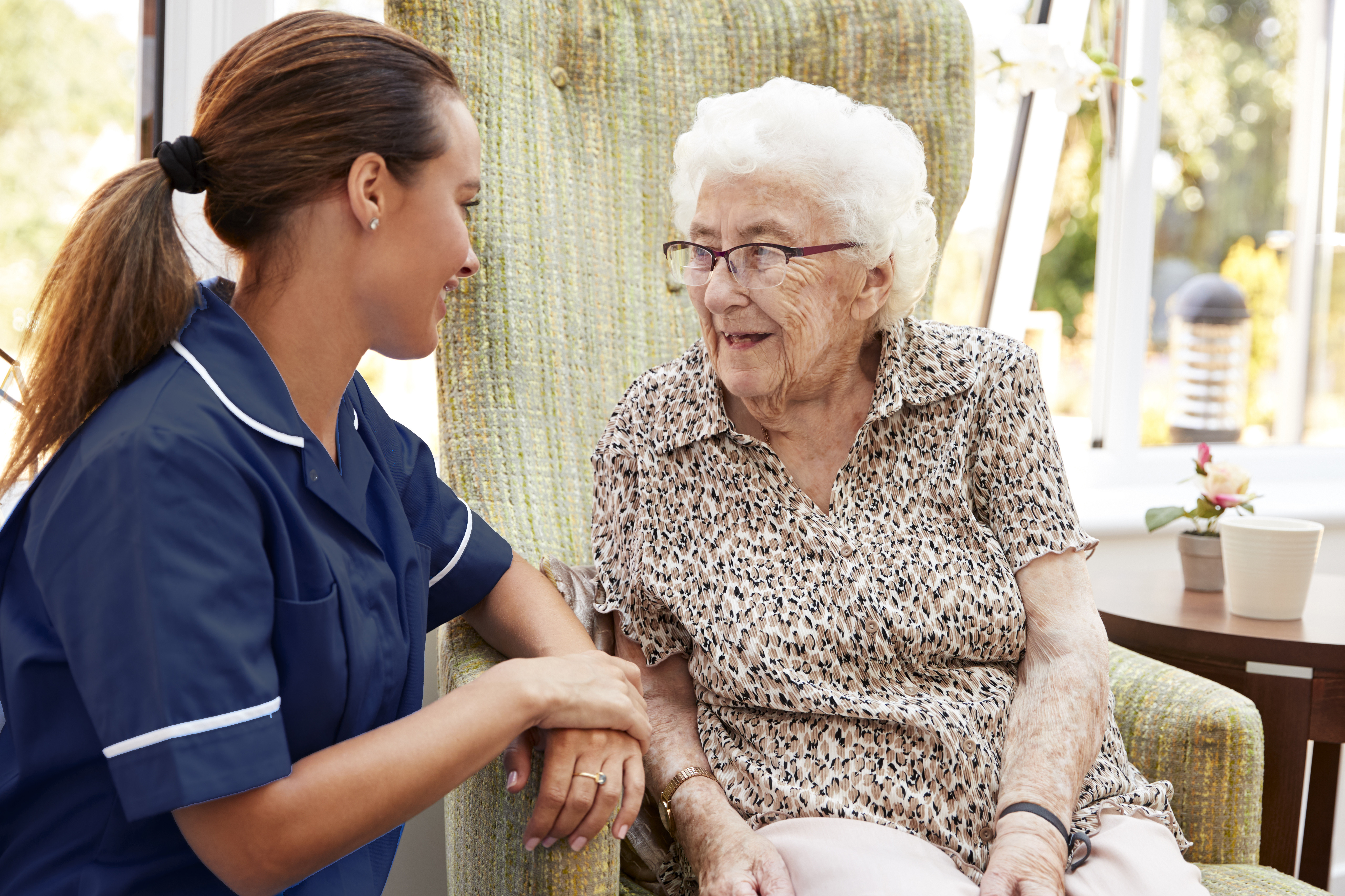 A nurse in a blue uniform is talking to an elderly woman who is sitting in a comfortable chair