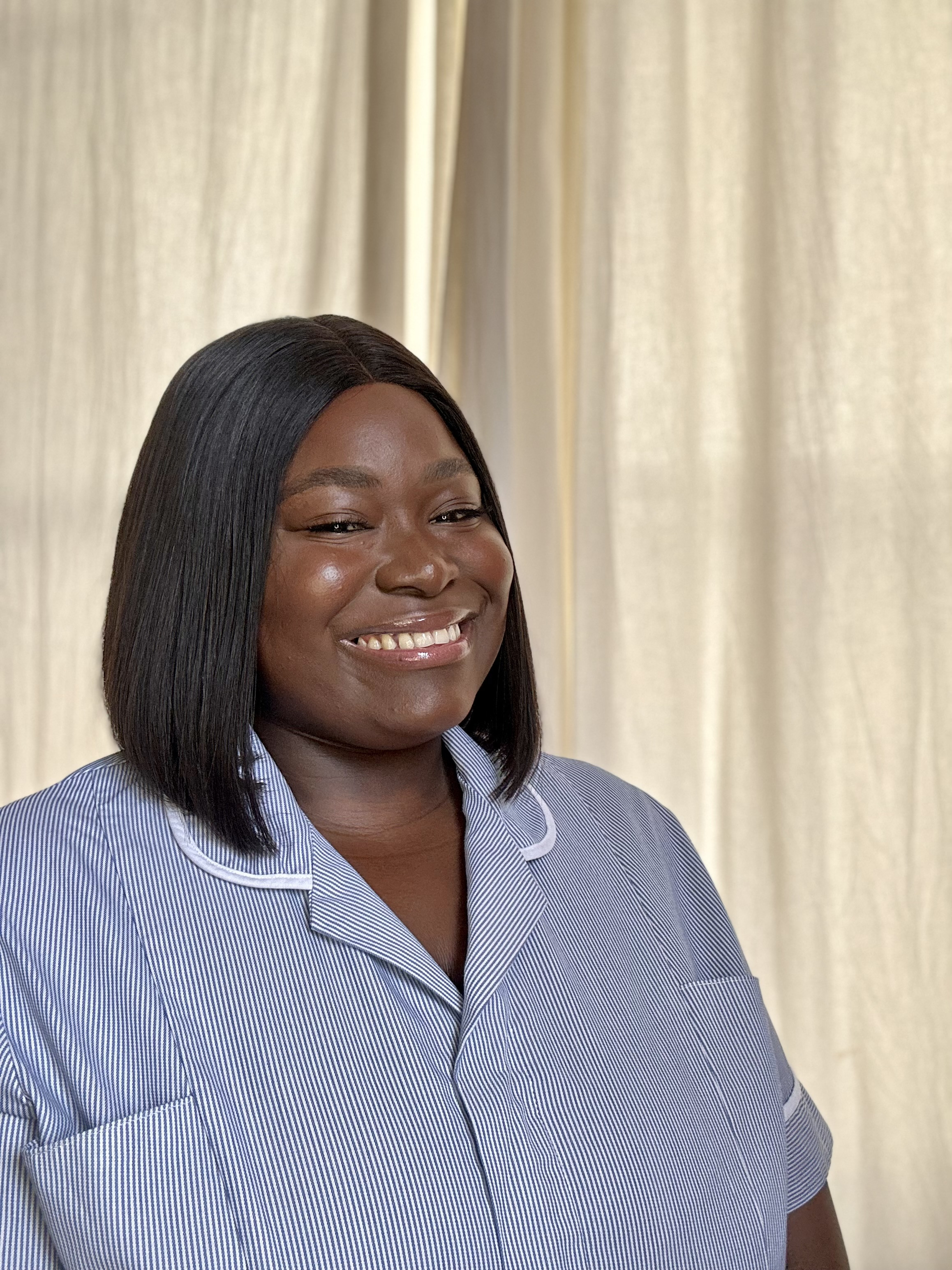 Yaggon Gwani. She is wearing a light blue nurse's uniform with white trim and standing in front of a window with closed curtains
